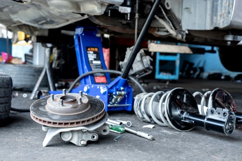 Various car parts including a brake disc, hub assembly, and MacPherson strut lie on a concrete garage floor next to a blue hydraulic floor jack.